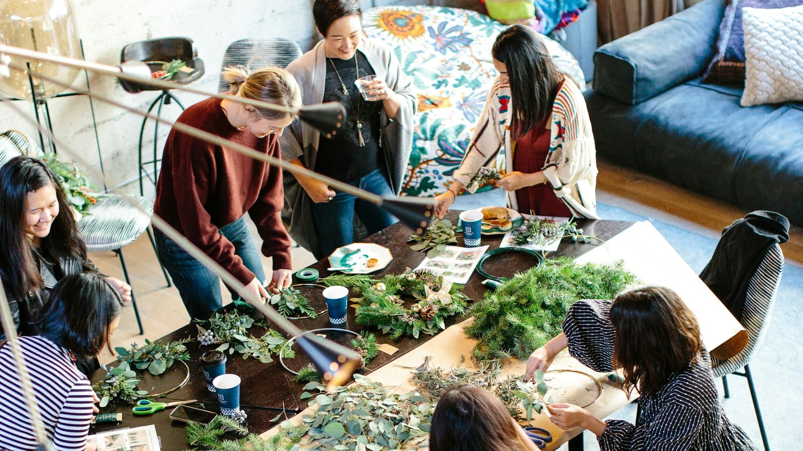Overhead view of a group of women making evergreen wreaths around a long workshop table covered in branches and eucalyptus