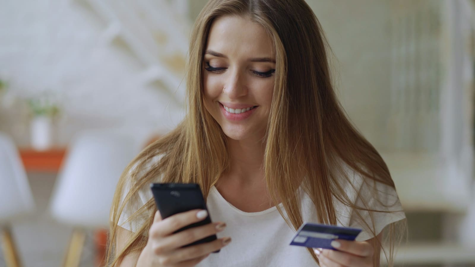 Smiling woman holding a smartphone in one hand and a bank card in the other while paying online