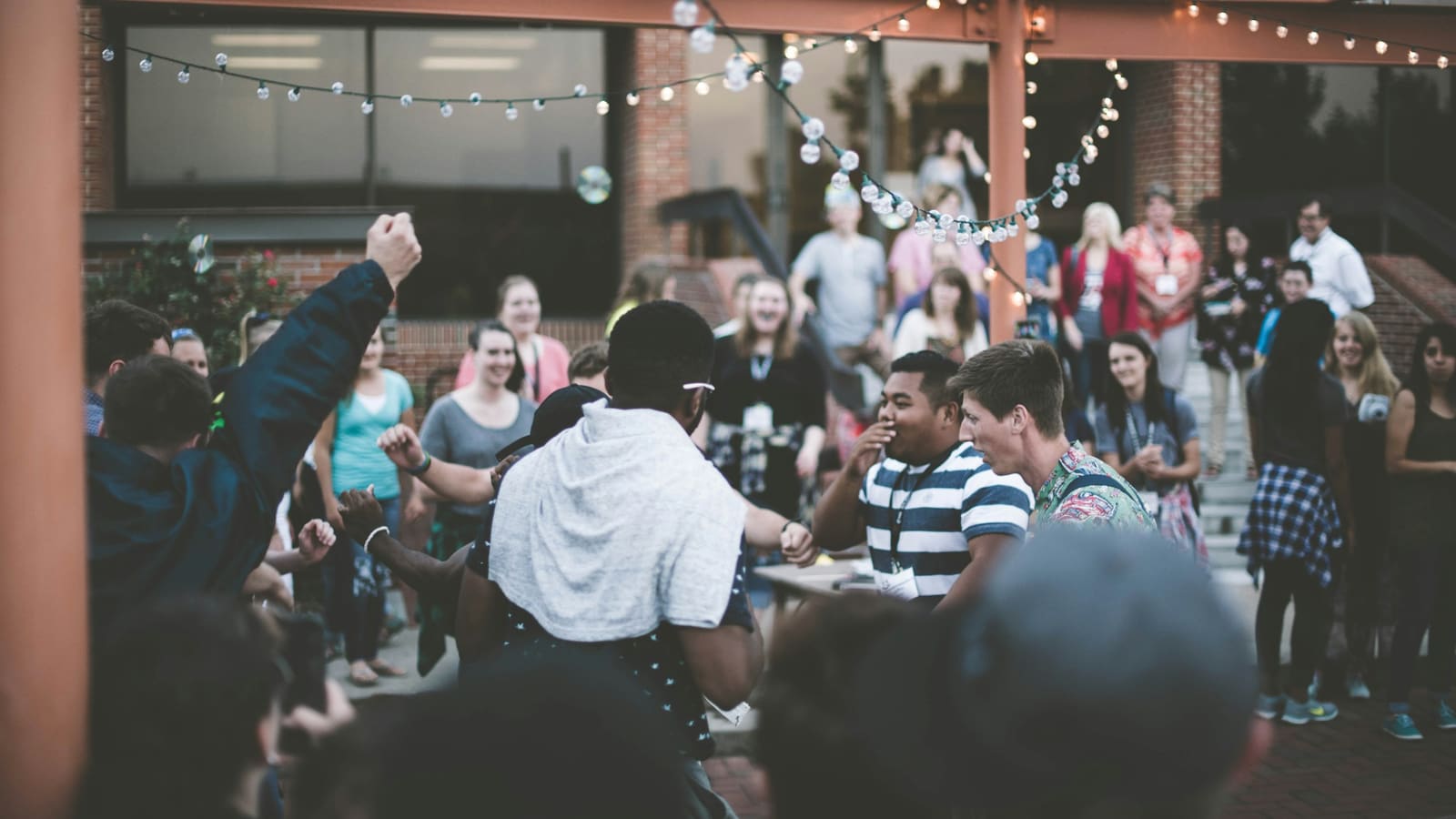 A crowd wearing name badges cheers on two people dancing at an outdoor evening gathering under strings of lights