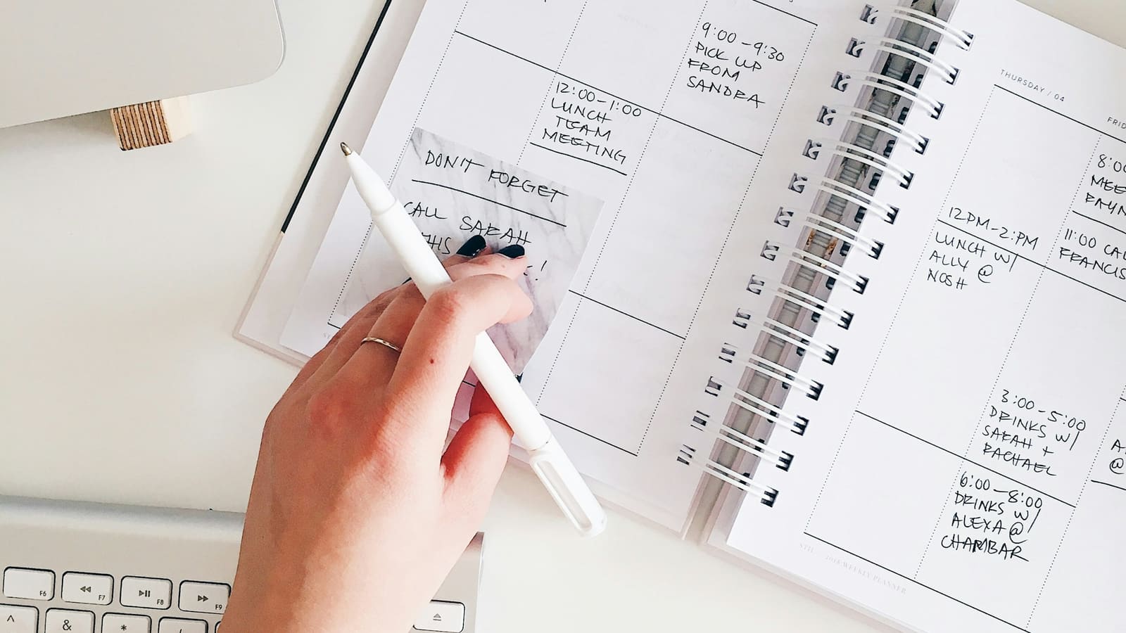 A hand holding a white pen over an open weekly planner filled with handwritten appointments, beside a computer keyboard