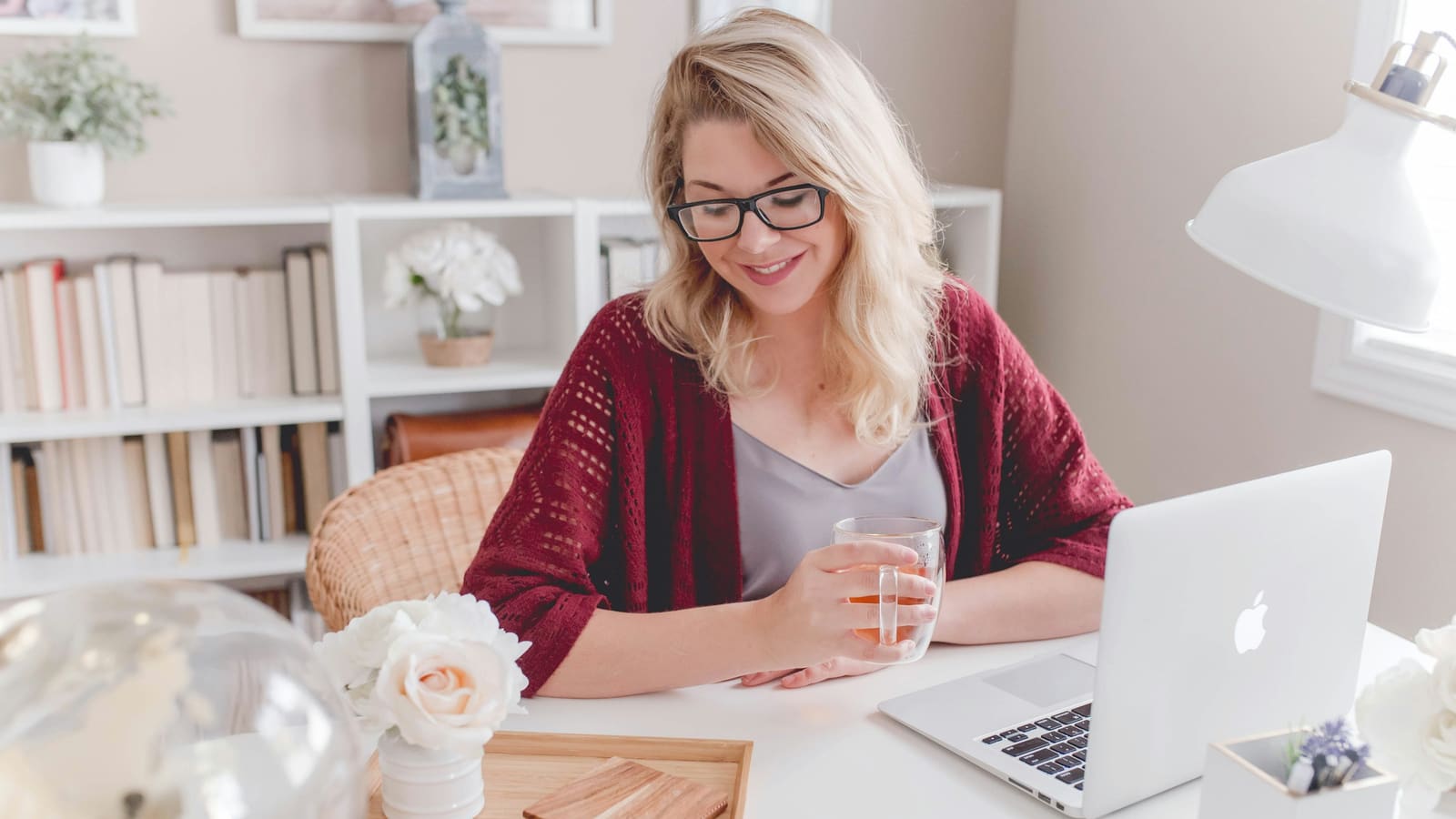 Smiling woman in glasses holding a glass mug of tea at a white desk with an open laptop and a vase of white roses