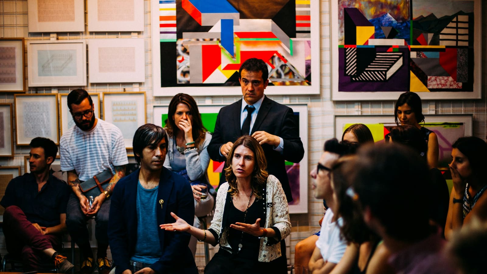 A small group seated and standing in an art gallery, listening to a woman speak during a discussion event